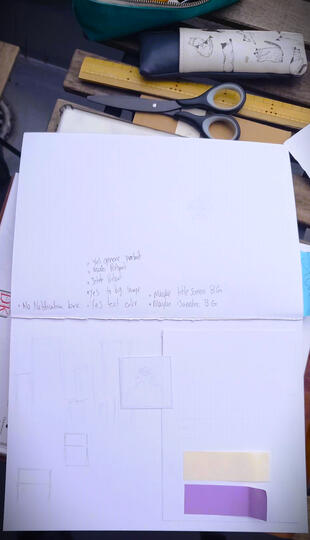 An outdoor picture of a white, open sketchbook with pencil notes and pieces of paper cut and layered on the page to mock up a game user interface and general appearance. Scissors and two pencil cases lie open nearby on a wooden picnic table.